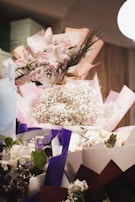 A collection of beautifully arranged flower bouquets featuring a variety of flowers including lilies, roses, and baby's breath. The bouquets are wrapped in colorful paper such as pink, purple, and white, and are placed against a softly lit background.
