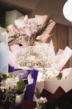 A collection of beautifully arranged flower bouquets featuring a variety of flowers including lilies, roses, and baby's breath. The bouquets are wrapped in colorful paper such as pink, purple, and white, and are placed against a softly lit background.