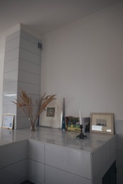 A minimalist interior corner features a ceramic tiled surface with decor items. Dried grass in a simple vase, framed artwork, and books are neatly arranged. An unlit white candle stands in a vintage holder, adding an artistic touch to the neutral space.