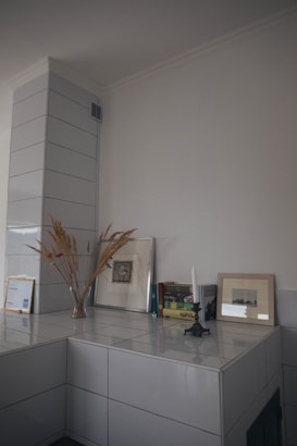 A minimalist interior corner features a ceramic tiled surface with decor items. Dried grass in a simple vase, framed artwork, and books are neatly arranged. An unlit white candle stands in a vintage holder, adding an artistic touch to the neutral space.