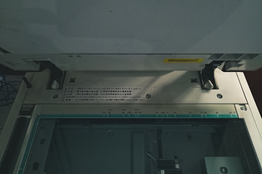 The image captures the interior of a copier or scanner machine. It shows the glass scanning bed with measurement markings for different paper sizes such as A3, A4, B5. There are cautionary labels in multiple languages on the machine's metallic edge. Two black latches and several screws are visible, indicating the functional components of the device. The lighting is dim, casting shadows inside the machinery.
