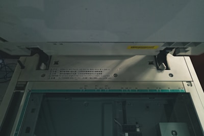 The image captures the interior of a copier or scanner machine. It shows the glass scanning bed with measurement markings for different paper sizes such as A3, A4, B5. There are cautionary labels in multiple languages on the machine's metallic edge. Two black latches and several screws are visible, indicating the functional components of the device. The lighting is dim, casting shadows inside the machinery.