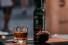 A glass of whiskey filled with ice cubes is placed on a wooden surface alongside a green bottle labeled Irish Whiskey. Next to the bottle is a small bowl containing peanuts. The background is blurred, giving focus to the whiskey, bottle, and nuts.