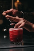 A pair of hands is seen delicately garnishing a vibrant red cocktail with a small spoon. The cocktail is placed on a reflective surface, and it appears to have a frothy top. The background is out of focus, creating a warm and intimate atmosphere.
