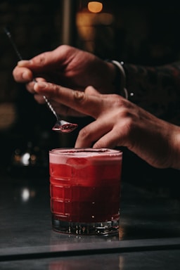 A pair of hands is seen delicately garnishing a vibrant red cocktail with a small spoon. The cocktail is placed on a reflective surface, and it appears to have a frothy top. The background is out of focus, creating a warm and intimate atmosphere.