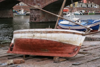 A small, worn-out boat is positioned on wooden planks by the water's edge, appearing to be undergoing repairs or maintenance. In the background, multiple boats are docked near a stone bridge with three arches. The weathered bridge and surrounding buildings suggest an old, historical waterfront area.