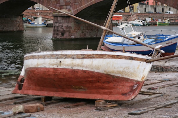 A small, worn-out boat is positioned on wooden planks by the water's edge, appearing to be undergoing repairs or maintenance. In the background, multiple boats are docked near a stone bridge with three arches. The weathered bridge and surrounding buildings suggest an old, historical waterfront area.