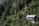 A small house with a metal roof is nestled among lush, densely packed trees on a forested hillside. The house has a balcony and is surrounded by vibrant greenery, with tall evergreen trees dominating the landscape.