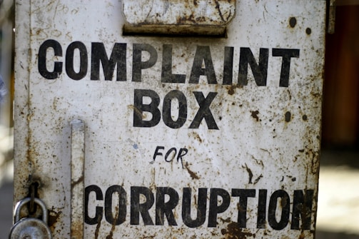 A rusty metal box with the words 'Complaint Box for Corruption' written in bold black letters. The surface is weathered and shows signs of age with patches of rust and dirt.