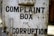 A rusty metal box with the words 'Complaint Box for Corruption' written in bold black letters. The surface is weathered and shows signs of age with patches of rust and dirt.