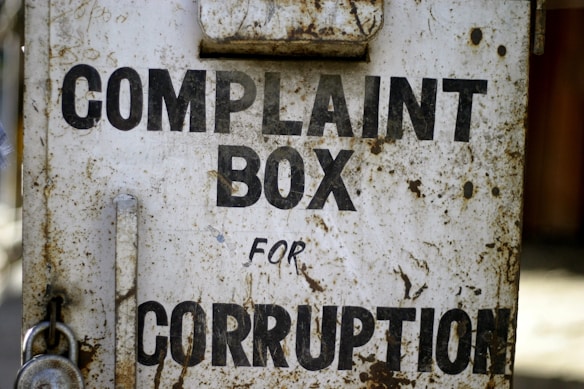 A rusty metal box with the words 'Complaint Box for Corruption' written in bold black letters. The surface is weathered and shows signs of age with patches of rust and dirt.