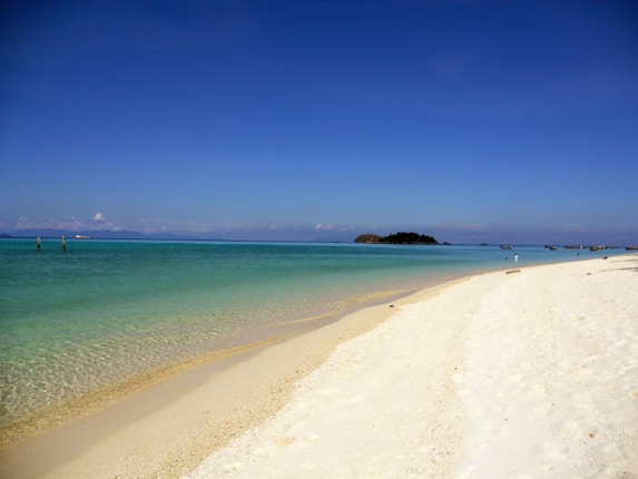 A serene beach view with clear blue skies, soft white sand, and calm turquoise water extending to the horizon. A small island is visible in the distance, adding to the tranquil and picturesque scenery.