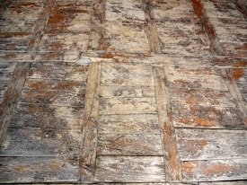 A close-up view of a worn and weathered wooden surface, characterized by its rough texture and visible grain patterns. The wood is arranged in a herringbone pattern and shows signs of age, with patches of peeling paint and exposed wood. The colors are muted and earthy, contributing to a rustic appearance.