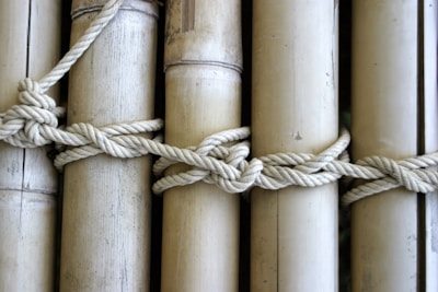A series of bamboo poles are tied together with a thick, knotted rope. The bamboo is smooth and light-colored, with visible natural texture and markings. The rope appears strong and woven tightly around the poles, emphasizing the craftsmanship of the knot.