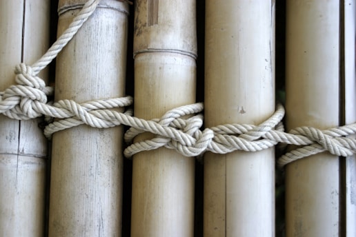 A series of bamboo poles are tied together with a thick, knotted rope. The bamboo is smooth and light-colored, with visible natural texture and markings. The rope appears strong and woven tightly around the poles, emphasizing the craftsmanship of the knot.