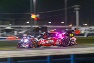 A sleek, high-performance racing car is captured in motion on a nighttime racetrack. The car is adorned with vibrant decals and a large number 53 on its side. Neon lights accentuate the vehicle, and bright headlights pierce through the darkness. The blurred background suggests high speed and includes stadium lights and possibly the outlines of a grandstand.
