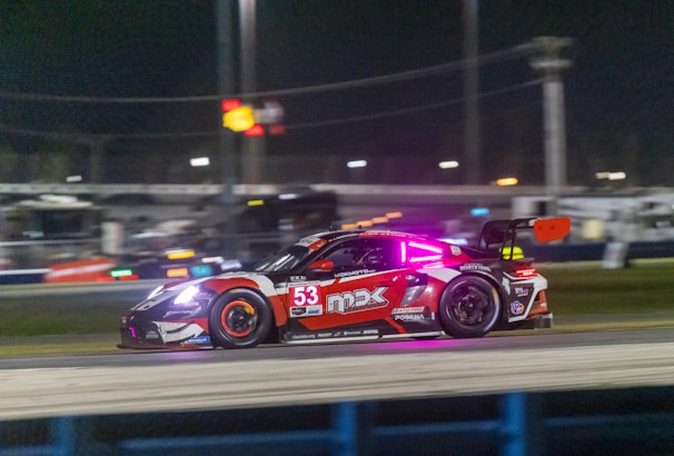 A sleek, high-performance racing car is captured in motion on a nighttime racetrack. The car is adorned with vibrant decals and a large number 53 on its side. Neon lights accentuate the vehicle, and bright headlights pierce through the darkness. The blurred background suggests high speed and includes stadium lights and possibly the outlines of a grandstand.