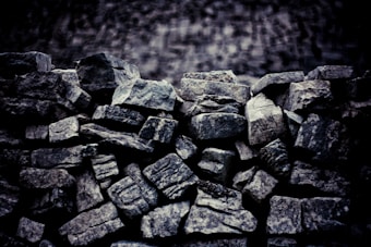 A close-up view of a pile of rough, irregularly shaped stones stacked together. The stones have varied textures and shades of gray, suggesting a natural and unprocessed state. The background is blurred, highlighting the rugged surfaces of the rocks.