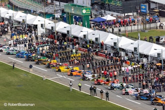 Aerial view of a motorsport event featuring a grid of colorful racing cars lined up in a row. Many people, including teams in uniforms and spectators, are gathered alongside tents and pit lane structures. The scene is bustling with activity, and there are various promotional signs and banners visible.