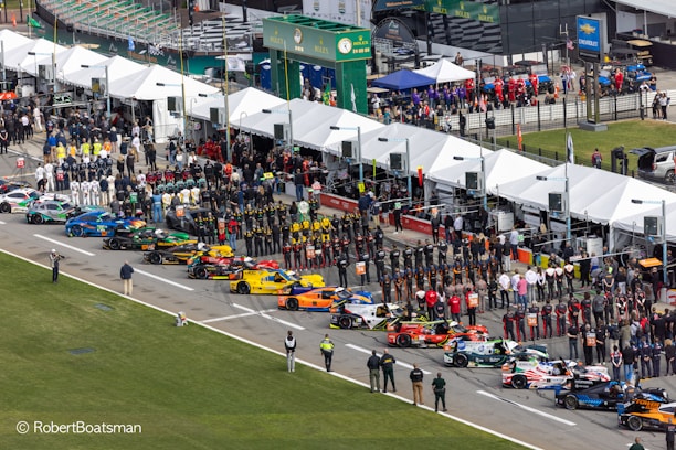 Aerial view of a motorsport event featuring a grid of colorful racing cars lined up in a row. Many people, including teams in uniforms and spectators, are gathered alongside tents and pit lane structures. The scene is bustling with activity, and there are various promotional signs and banners visible.