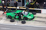 A vibrant green racing car is in a pit stop area, surrounded by several pit crew members in black uniforms and helmets. The crew is engaged in activities like changing tires and refueling. The car features various sponsor logos and has the number 63 on its side. In the background, additional crew members and equipment are visible.