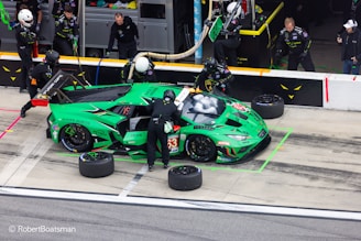 A vibrant green racing car is in a pit stop area, surrounded by several pit crew members in black uniforms and helmets. The crew is engaged in activities like changing tires and refueling. The car features various sponsor logos and has the number 63 on its side. In the background, additional crew members and equipment are visible.