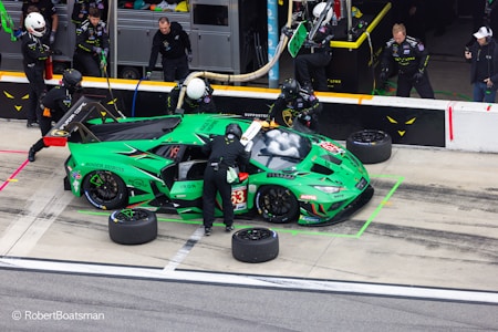 A vibrant green racing car is in a pit stop area, surrounded by several pit crew members in black uniforms and helmets. The crew is engaged in activities like changing tires and refueling. The car features various sponsor logos and has the number 63 on its side. In the background, additional crew members and equipment are visible.