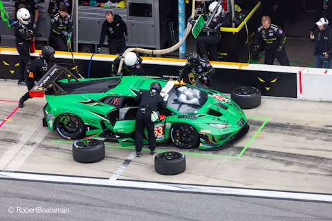 A vibrant green racing car is in a pit stop area, surrounded by several pit crew members in black uniforms and helmets. The crew is engaged in activities like changing tires and refueling. The car features various sponsor logos and has the number 63 on its side. In the background, additional crew members and equipment are visible.