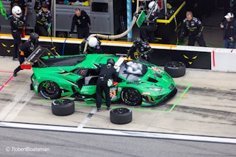 A vibrant green racing car is in a pit stop area, surrounded by several pit crew members in black uniforms and helmets. The crew is engaged in activities like changing tires and refueling. The car features various sponsor logos and has the number 63 on its side. In the background, additional crew members and equipment are visible.