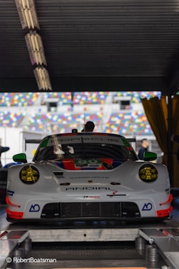 A white racing car with green and red accents is positioned in a racing garage. The car features prominent logos and branding, including 'Porsche' on the windshield. Behind it, colorful, out-of-focus seating is visible through an open garage door.