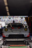A high-performance Porsche racing car is positioned in a workshop or garage area. The car has a sleek, aerodynamic design with prominent headlights and a detailed front bumper. It features a number of sponsors' logos on its body and is parked on a platform with various technical equipment around. There are multicolored seating sections of a racing track visible through the open area behind the car.