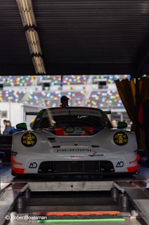 A high-performance Porsche racing car is positioned in a workshop or garage area. The car has a sleek, aerodynamic design with prominent headlights and a detailed front bumper. It features a number of sponsors' logos on its body and is parked on a platform with various technical equipment around. There are multicolored seating sections of a racing track visible through the open area behind the car.