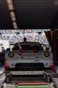 A high-performance Porsche racing car is positioned in a workshop or garage area. The car has a sleek, aerodynamic design with prominent headlights and a detailed front bumper. It features a number of sponsors' logos on its body and is parked on a platform with various technical equipment around. There are multicolored seating sections of a racing track visible through the open area behind the car.