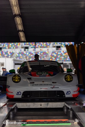 A high-performance Porsche racing car is positioned in a workshop or garage area. The car has a sleek, aerodynamic design with prominent headlights and a detailed front bumper. It features a number of sponsors' logos on its body and is parked on a platform with various technical equipment around. There are multicolored seating sections of a racing track visible through the open area behind the car.
