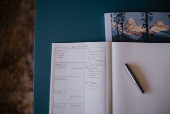 An open planner or notebook lies on a table. The page is divided into sections for different days of the week, with handwritten notes and tasks detailing a weekly schedule. A pen rests on the right side of the open page. The upper portion of the image shows a closed book with an illustration of mountain scenery.