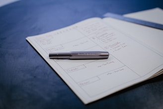 An open notebook contains handwritten notes organized in a grid layout. A pen rests on top of the notebook, and part of a metallic object, possibly a ruler or bookmark, is visible. The surface beneath is a smooth, blue material.