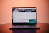 A laptop is placed on a table with its screen displaying a dashboard interface featuring a calendar and various navigation options. The background is a soft gradient from pink to orange, adding a warm ambiance to the scene.