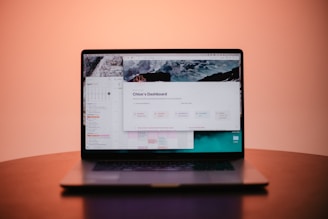 A laptop is placed on a table with its screen displaying a dashboard interface featuring a calendar and various navigation options. The background is a soft gradient from pink to orange, adding a warm ambiance to the scene.