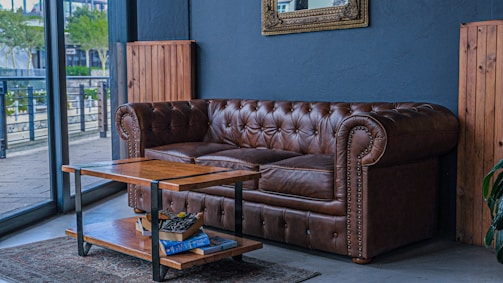 A cozy living room area featuring a classic brown leather Chesterfield sofa placed against a dark blue wall. A wooden coffee table with a metal frame stands in front of the sofa, holding several books and a magazine. A large decorative mirror is mounted above the sofa. The space is adjacent to a large window, letting in natural light and providing a view of the outside. Wooden paneling and decor elements contribute to a warm and rustic ambience.