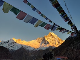 A stunning mountain landscape with a peak illuminated by golden sunlight, surrounded by snow-covered slopes. Multicolored prayer flags are strung across the foreground, adding a touch of cultural significance. Below, silhouettes of people are visible, gazing towards the mountain.
