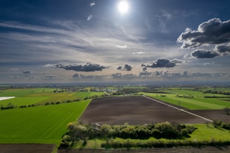 A vast agricultural landscape stretches out under a partly cloudy sky with the sun shining brightly. The fields are divided into sections of green crops and dark, freshly tilled soil. In the distance, a line of trees and small structures can be seen along the horizon.