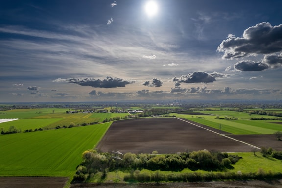 A vast agricultural landscape stretches out under a partly cloudy sky with the sun shining brightly. The fields are divided into sections of green crops and dark, freshly tilled soil. In the distance, a line of trees and small structures can be seen along the horizon.