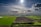 A vast agricultural landscape stretches out under a partly cloudy sky with the sun shining brightly. The fields are divided into sections of green crops and dark, freshly tilled soil. In the distance, a line of trees and small structures can be seen along the horizon.