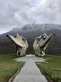 A monumental, abstract stone sculpture with sharp, angular forms stands in a grassy field, framed by a pathway leading towards it. Behind the sculpture, there is a backdrop of mountains partly covered with snow and a cloudy sky, creating a dramatic and awe-inspiring landscape.