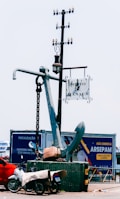 A large anchor stands upright on a platform, with a metal chain attached. A nearby pole supports a hanging street lamp and a sign reading 'Porto de Manaus.' In the foreground, a motorcycle covered with a tarp is parked next to the platform. Behind the anchor, a billboard displays advertising in Portuguese.