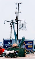A large anchor stands upright on a platform, with a metal chain attached. A nearby pole supports a hanging street lamp and a sign reading 'Porto de Manaus.' In the foreground, a motorcycle covered with a tarp is parked next to the platform. Behind the anchor, a billboard displays advertising in Portuguese.