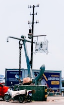A large anchor stands upright on a platform, with a metal chain attached. A nearby pole supports a hanging street lamp and a sign reading 'Porto de Manaus.' In the foreground, a motorcycle covered with a tarp is parked next to the platform. Behind the anchor, a billboard displays advertising in Portuguese.