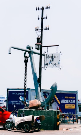 A large anchor stands upright on a platform, with a metal chain attached. A nearby pole supports a hanging street lamp and a sign reading 'Porto de Manaus.' In the foreground, a motorcycle covered with a tarp is parked next to the platform. Behind the anchor, a billboard displays advertising in Portuguese.
