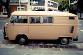 A beige vintage van is parked on the side of a street next to a sidewalk. The surroundings include trees and a red brick building with white trim and signage. In the background, a shop with an awning and various signs is visible.