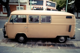 A beige vintage van is parked on the side of a street next to a sidewalk. The surroundings include trees and a red brick building with white trim and signage. In the background, a shop with an awning and various signs is visible.
